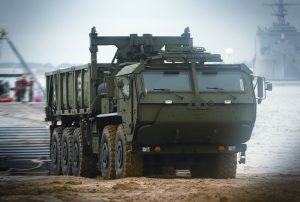 A large military truck with eight wheels drives on sandy terrain near water, carrying cargo in an open-bed container. The vehicle is dark green and has a crane arm on top, with blurred ships and personnel in the background.