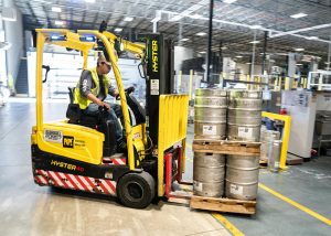 A worker drives a yellow forklift carrying several metal beer kegs on a wooden pallet inside a large, well-lit warehouse with high ceilings and industrial equipment.