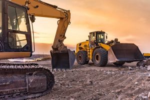 Two large yellow construction vehicles, an excavator and a front loader, are parked on a dirt construction site at sunset, with rocky soil in the foreground and a colorful sky in the background.
