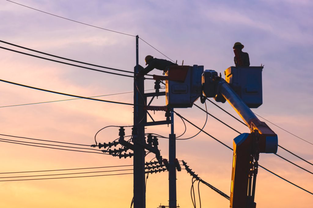 Two utility workers in raised buckets repair power lines on poles at sunset, silhouetted against a colorful sky.