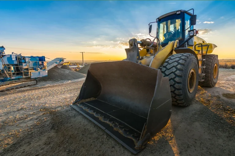 A large yellow front loader construction vehicle is parked on a dirt site at sunset, with its wide bucket facing forward and machinery visible in the background.